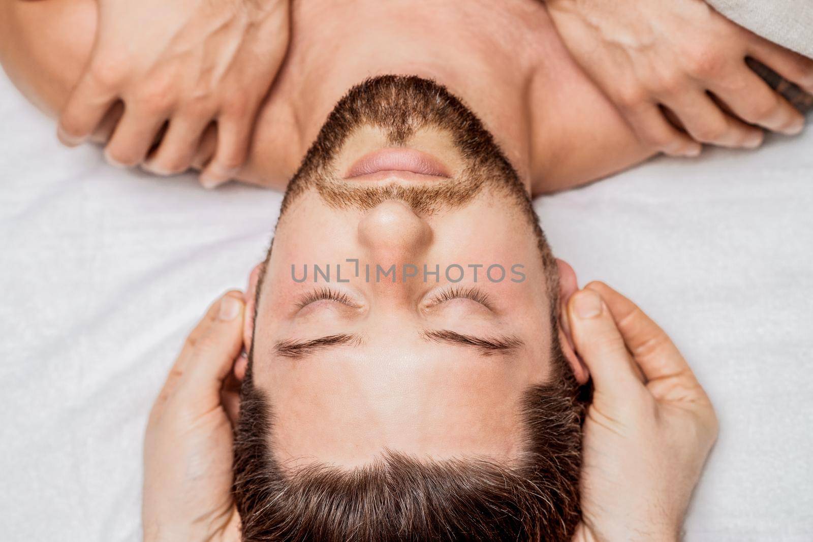 Handsome young man receiving relaxing head massage from a two masseurs in four hands in a health spa center