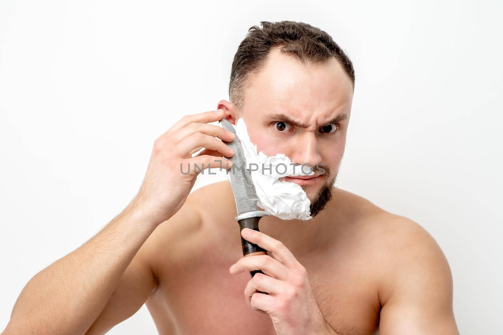Young caucasian man shaving beard with knife on white background