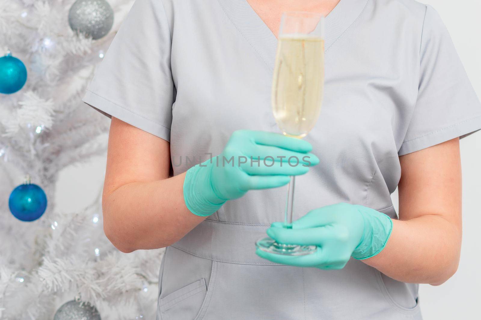 Beautiful young female doctor in protective gloves with glass of champagne standing near the christmas tree. Defocused