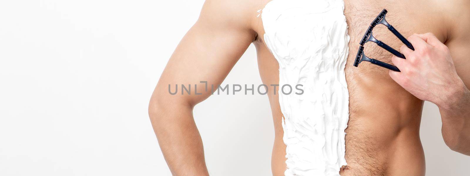 Young caucasian man with beard holds razor shaves his chest with white shaving foam on white background. Man shaving his torso