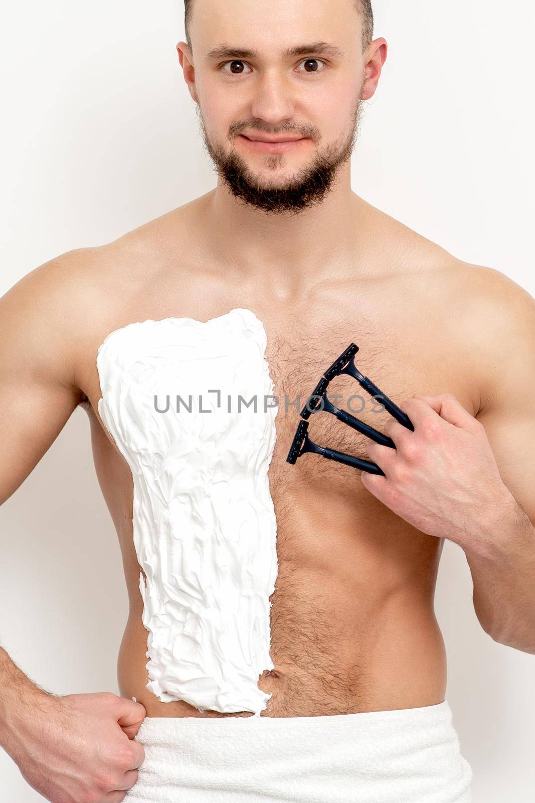 Young caucasian man with beard holds razor shaves his chest with white shaving foam on white background. Man shaving his torso