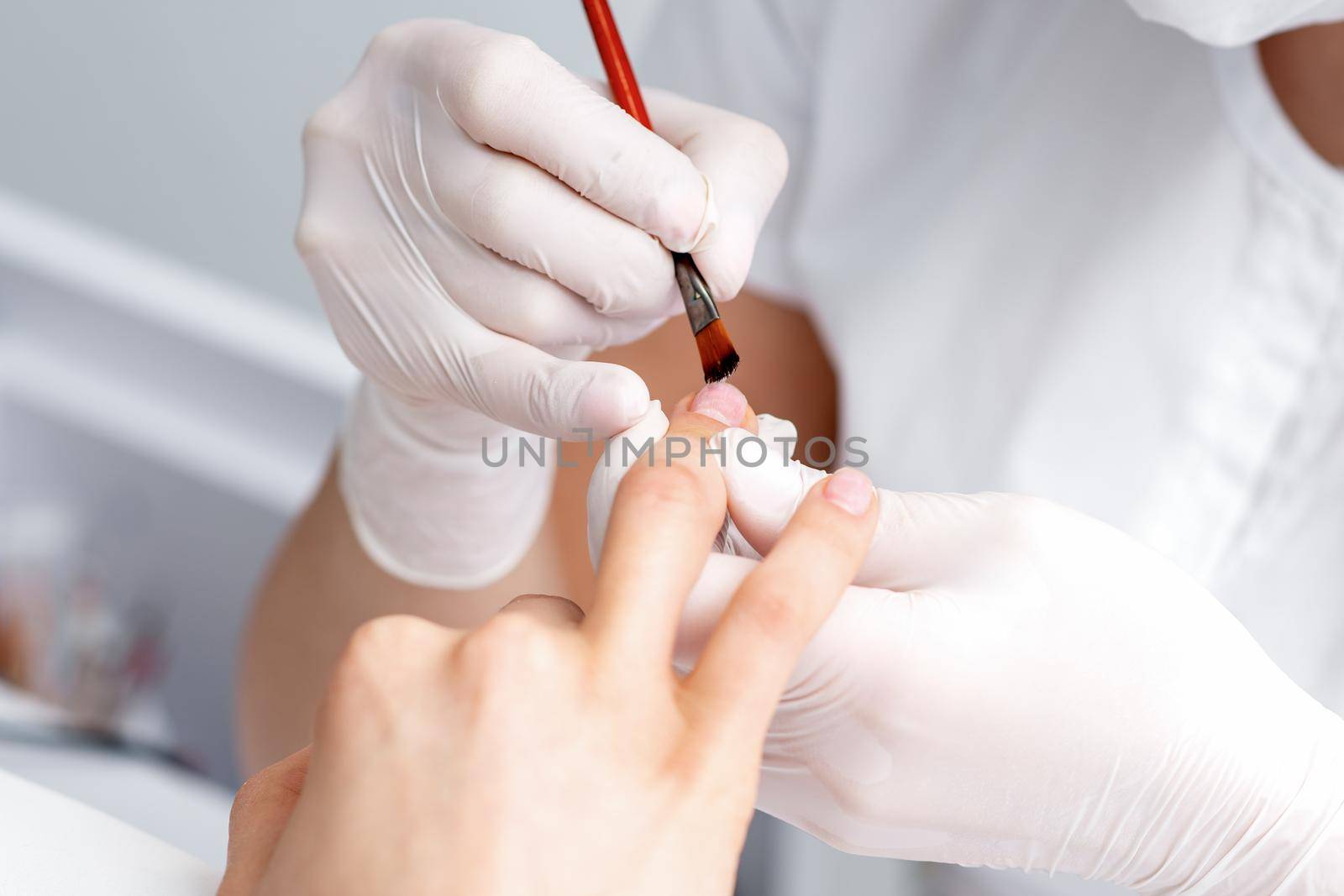 Manicure master in rubber gloves applying transparent nail polish on female nails in beauty salon