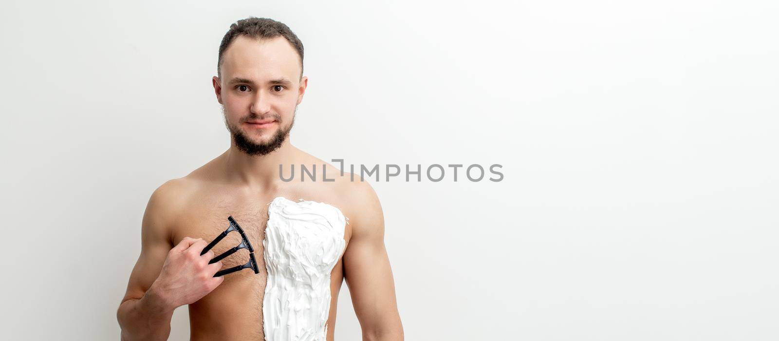 Young caucasian man with beard holds razor shaves his chest with white shaving foam on white background. Man shaving his torso