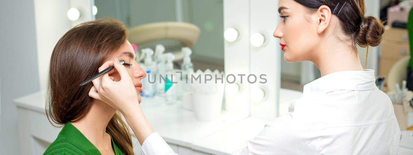 Makeup artist applies eyeshadow powder on eyes of young caucasian woman by brush tool in beauty salon