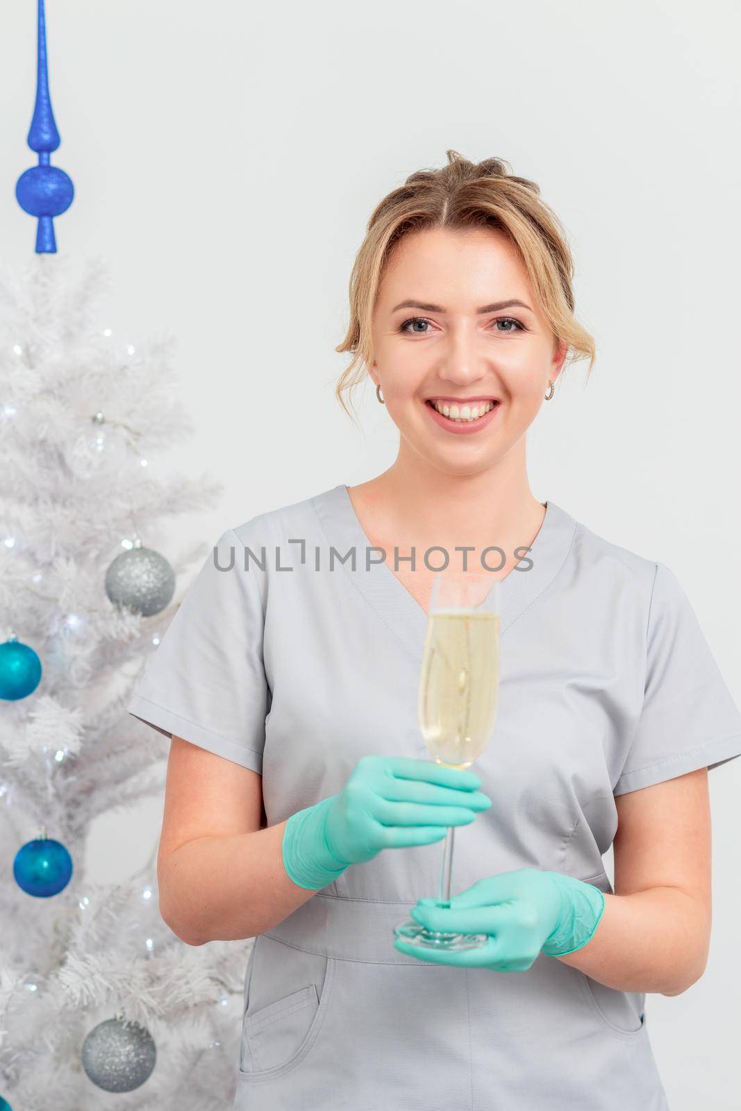 Beautiful young caucasian smiling female doctor in protective gloves with glass of champagne standing near the christmas tree