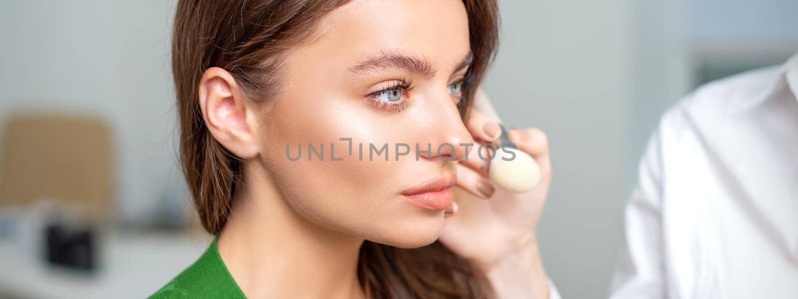 Makeup artist applying dry cosmetic tonal foundation on the face of young woman using brush tool in beauty salon