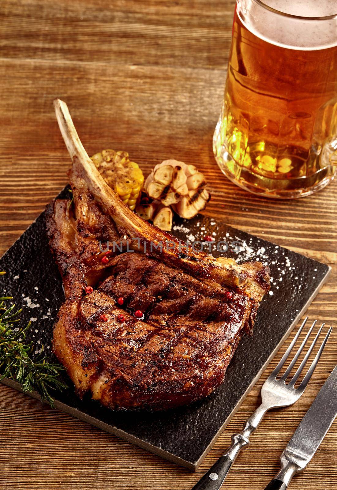 Grilled cowboy beef steak, glass of beer, herbs and spices on a wooden background. Top view. Still life