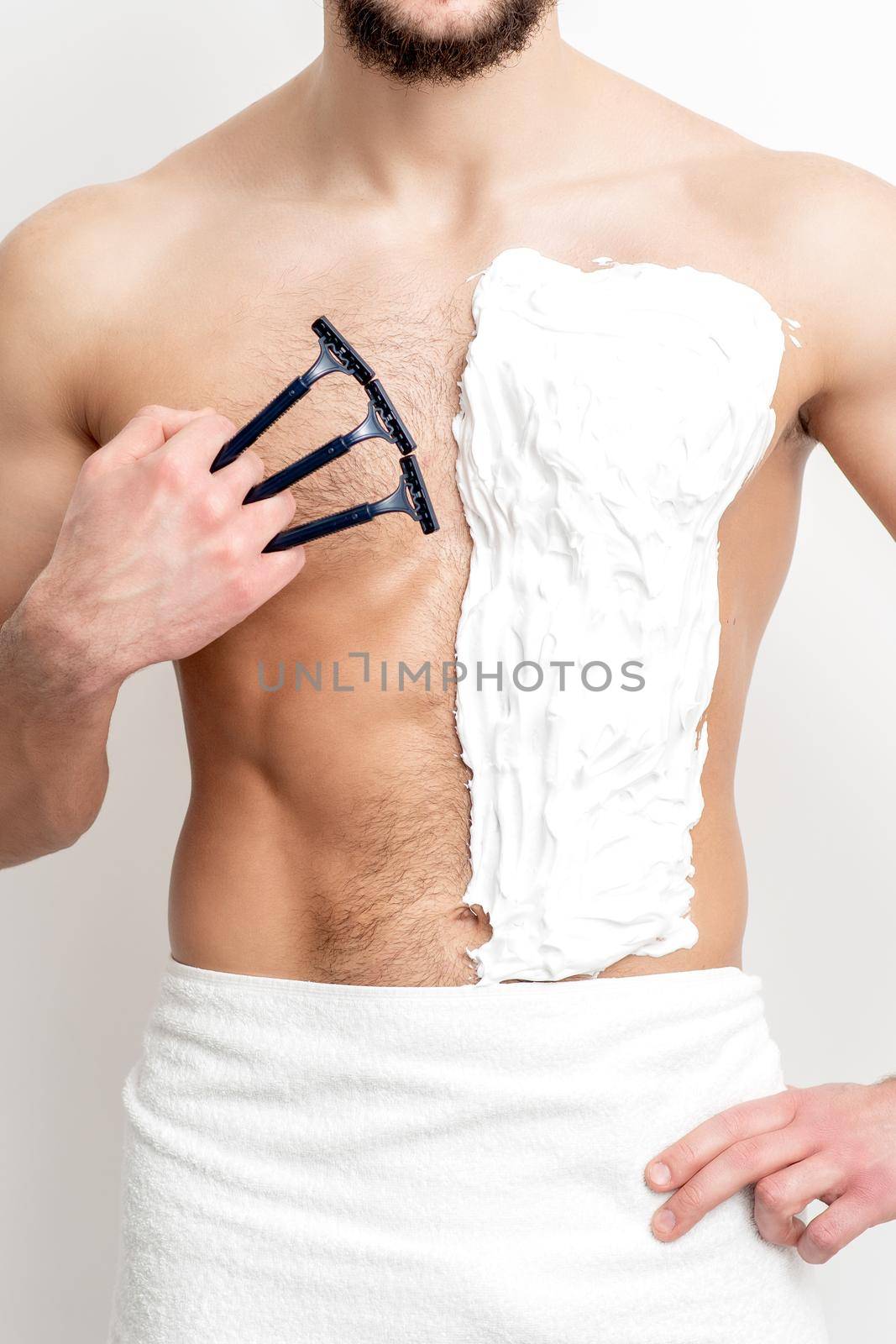 Young caucasian man with beard holds razor shaves his chest with white shaving foam on white background. Man shaving his torso