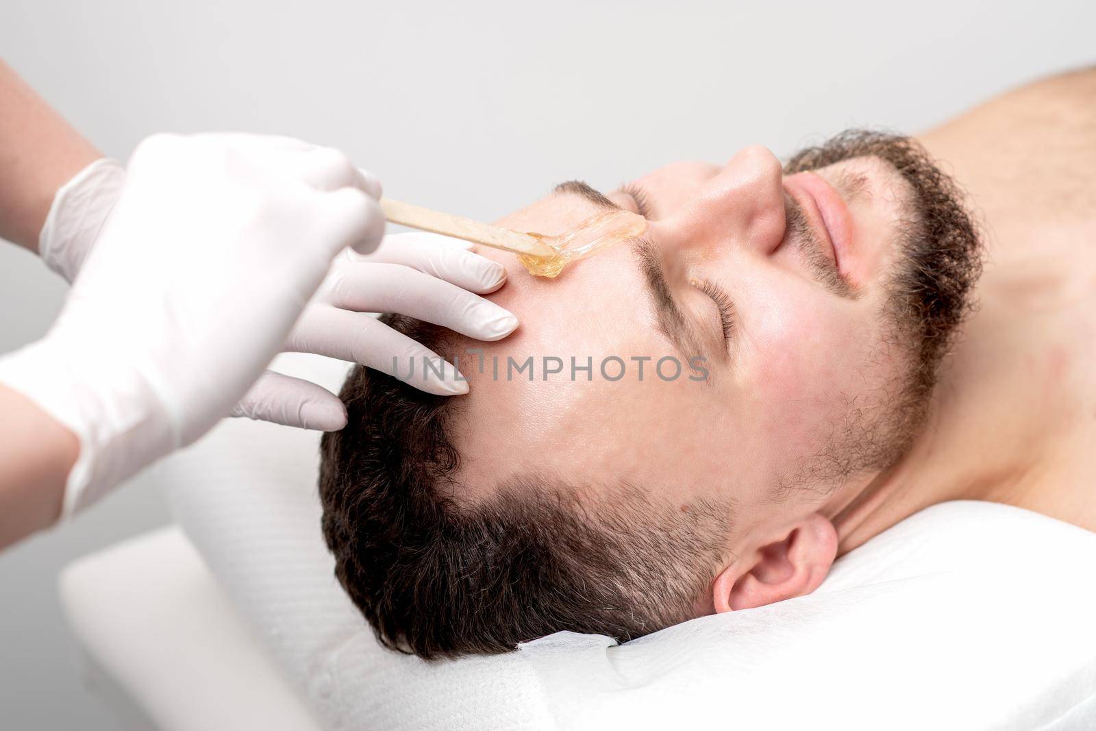 Beautician applies wax between male eyebrows before the procedure of waxing in the beauty salon