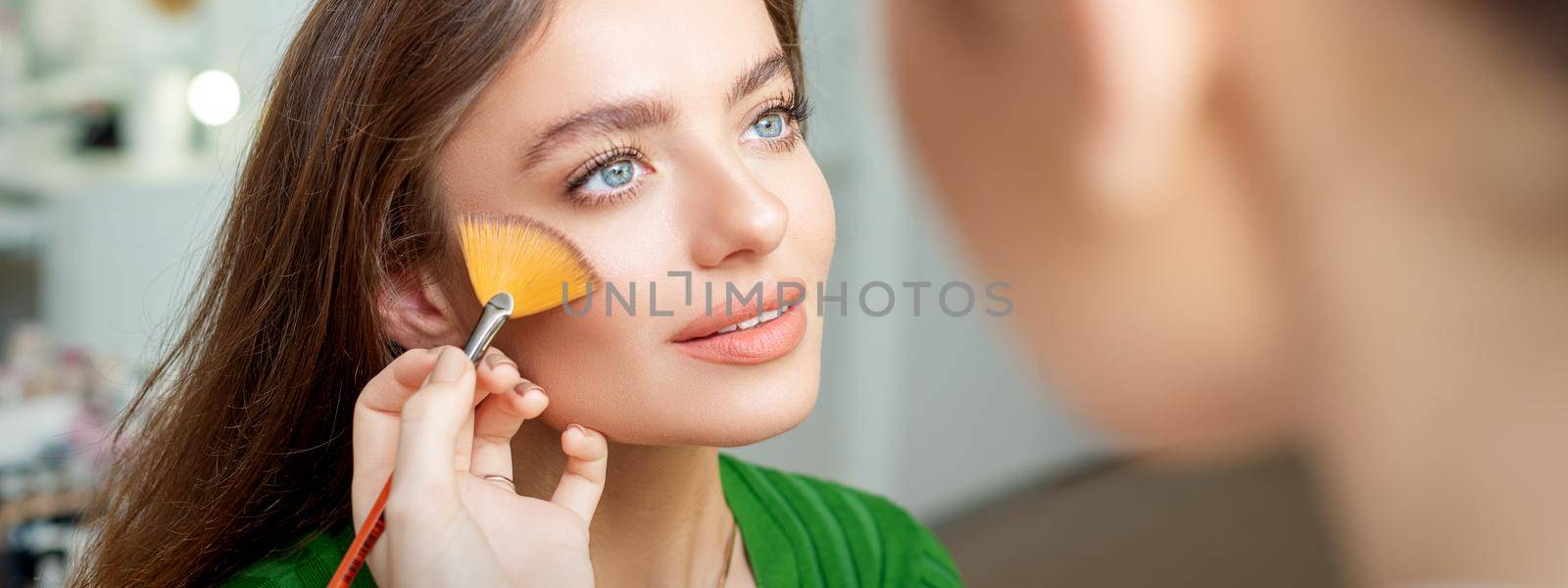 Professional make up artist applying powder by brush on cheeks of beautiful young caucasian woman in beauty salon