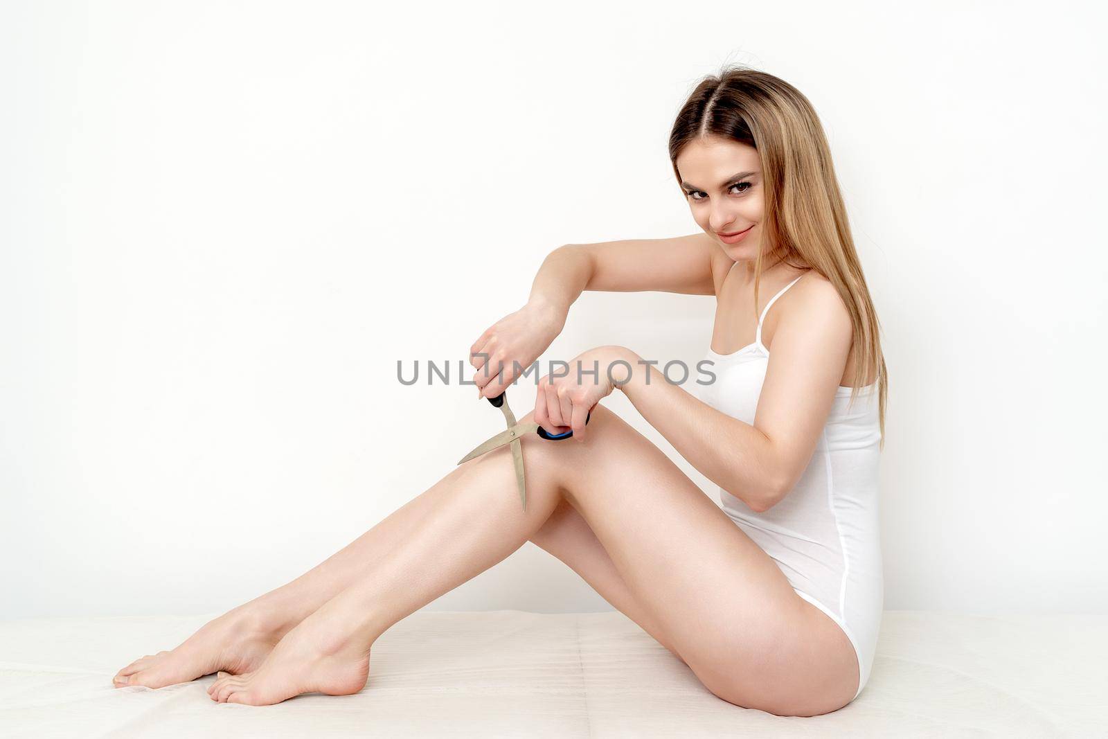 Beautiful young slim caucasian woman cutting hair on her legs by scissors on white background. Depilation concept