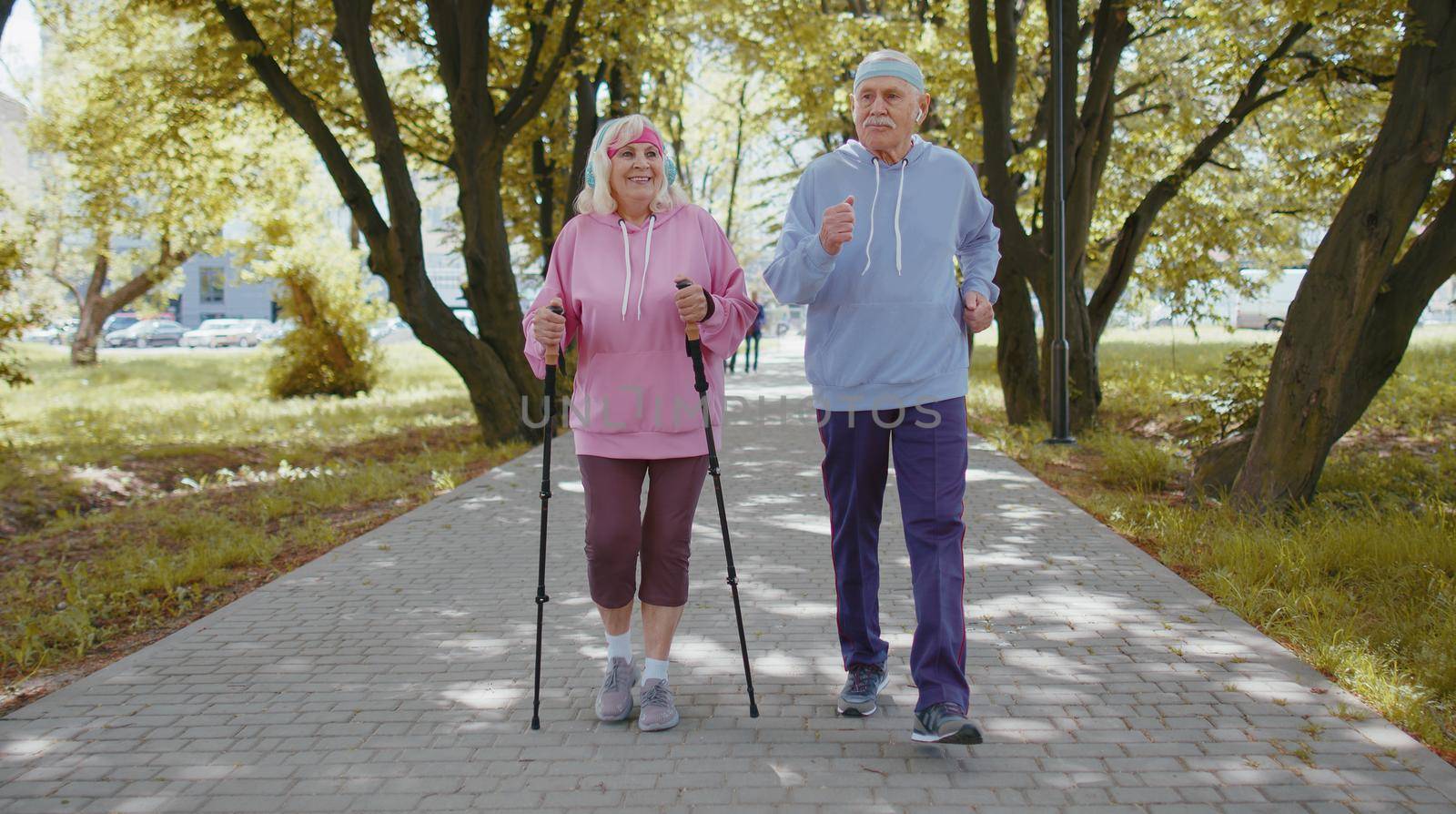 Active senior elderly couple 80 years old in summer park. Caucasian grandfather man training Nordic walking with ski trekking poles. Grandmother woman is running. Mature retired family sport hobby