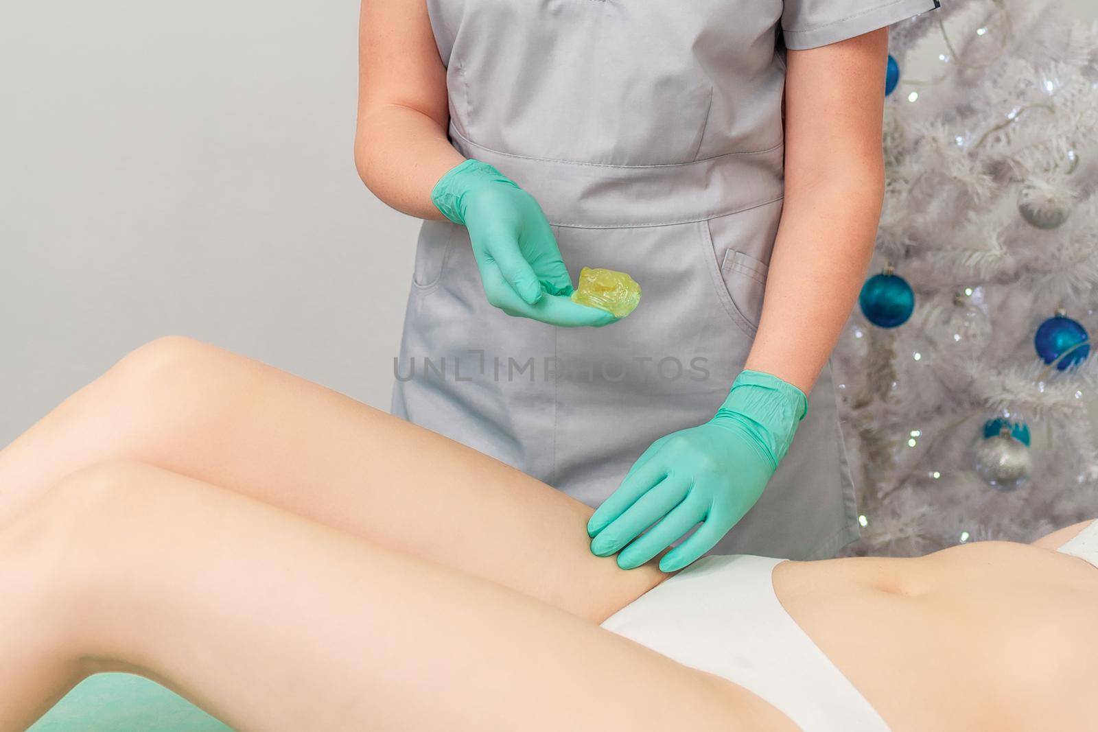 Cosmetologist in gloves applying paste for sugaring depilation on legs of young woman. Christmas discount waxing