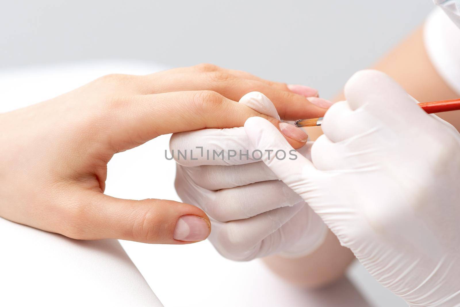 Manicure master in rubber gloves applying transparent nail polish on female nails in beauty salon