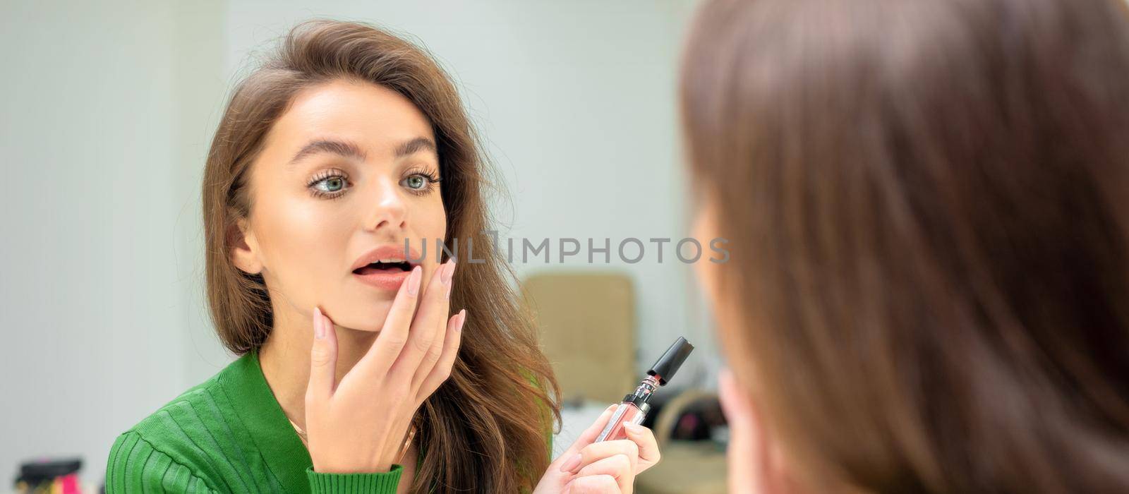 Beautiful young caucasian woman applies lipstick on her lips with her finger looking in mirror