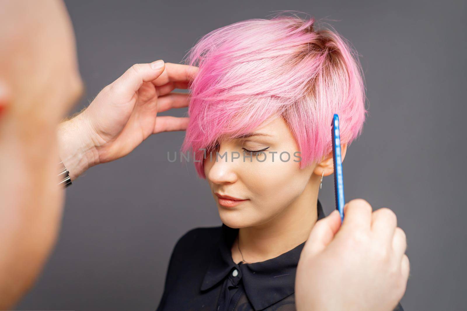 Hairdresser checking short pink hairstyle of young woman on gray background
