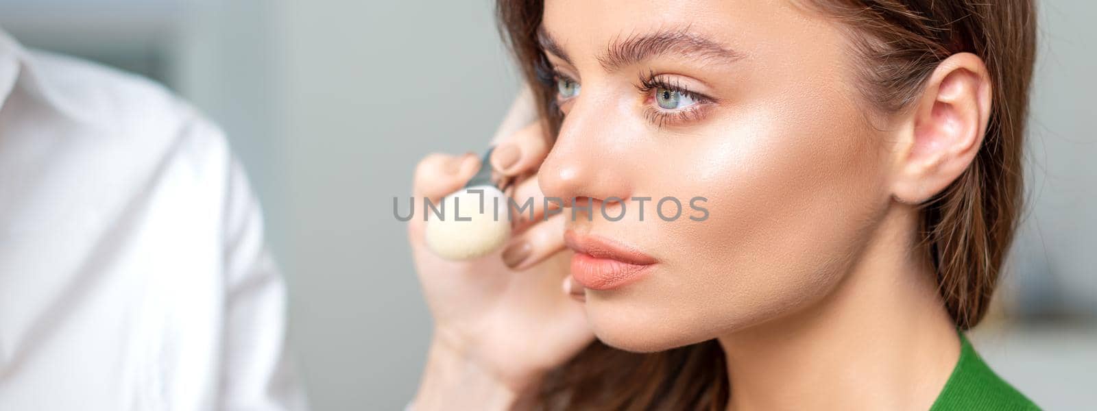 Makeup artist applying dry cosmetic tonal foundation on the face of young woman using brush tool in beauty salon