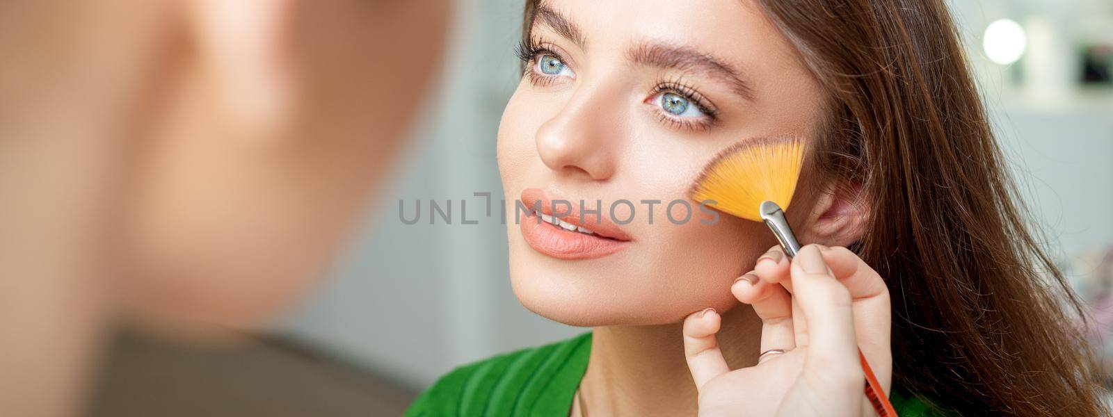 Professional make up artist applying powder by brush on cheeks of beautiful young caucasian woman in beauty salon