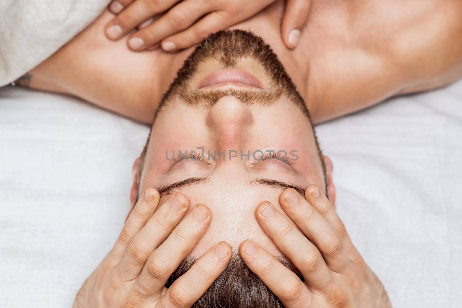 Handsome young man receiving relaxing head massage by four hand of two masseurs in health spa center
