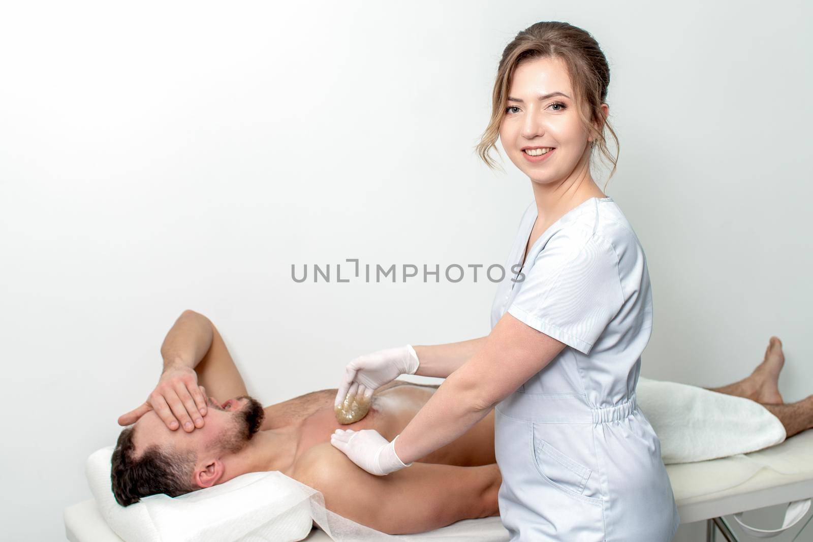 Young man receiving waxing chest by young female cosmetologist in beauty salon. Portrait of young female cosmetologist during waxing male chest
