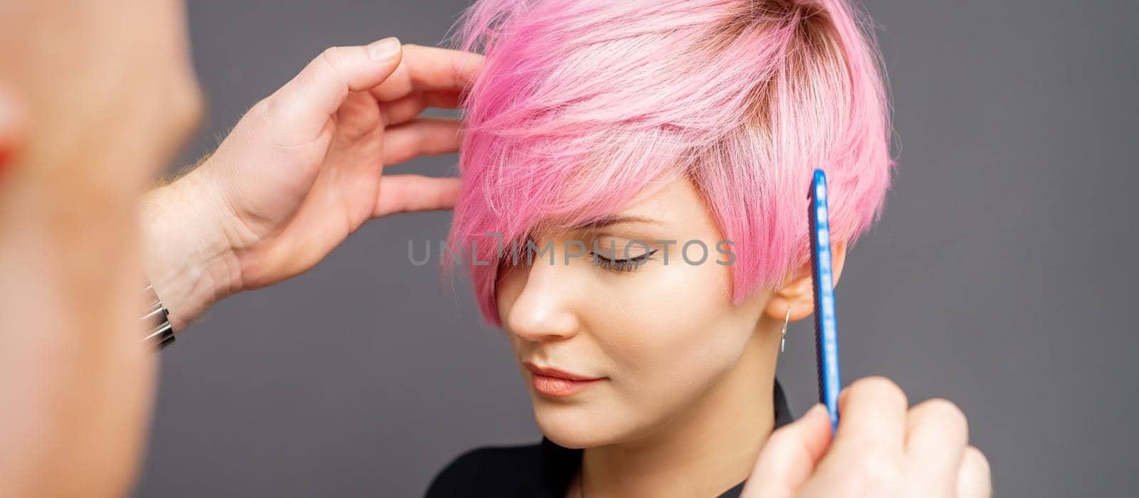 Hairdresser checking short pink hairstyle of young woman on gray background