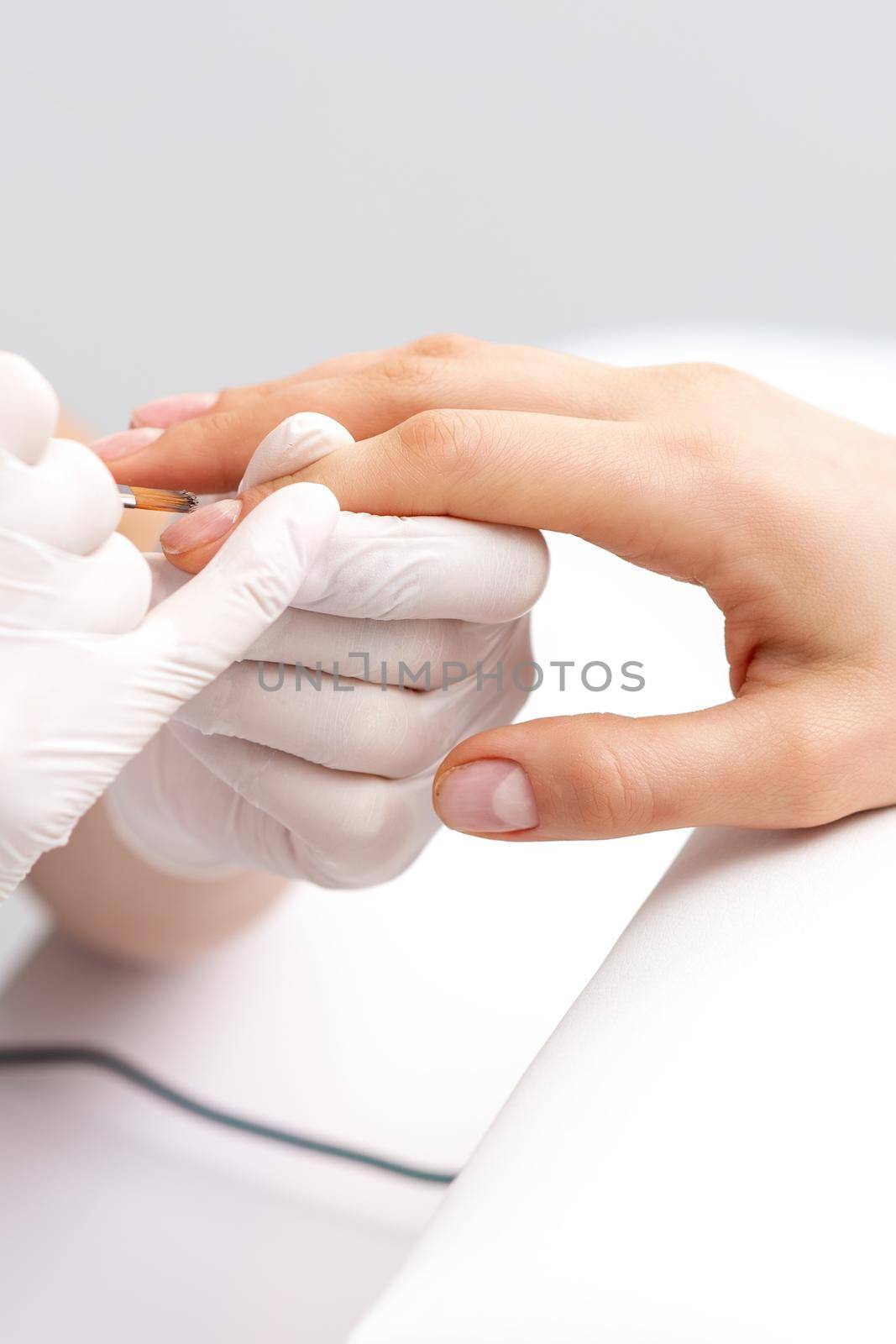 Manicure master in rubber gloves applying transparent nail polish on female nails in beauty salon