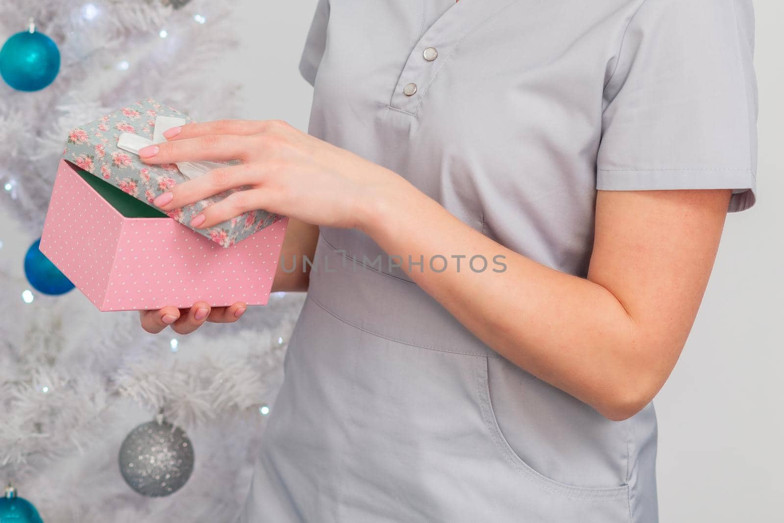 Beautiful young caucasian woman smiling in medical uniform with gift box near christmas tree on light background