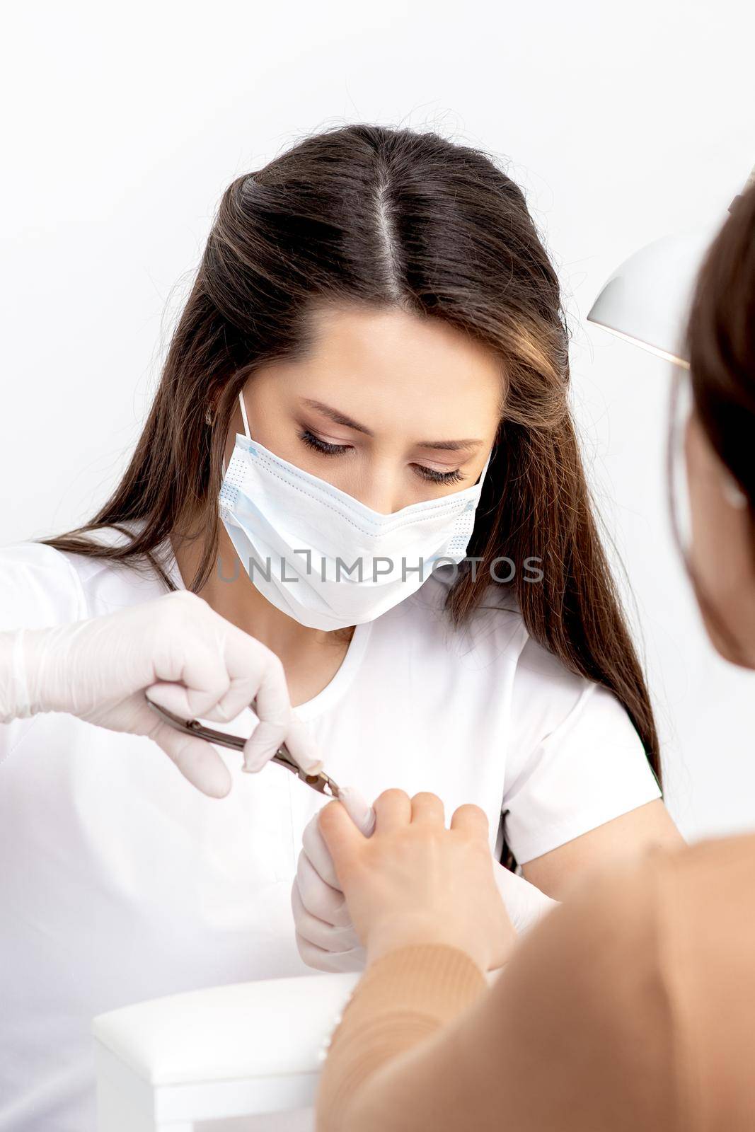 Manicurist in protective mask using manicure cutter to remove cuticle of female nails in manicure salon