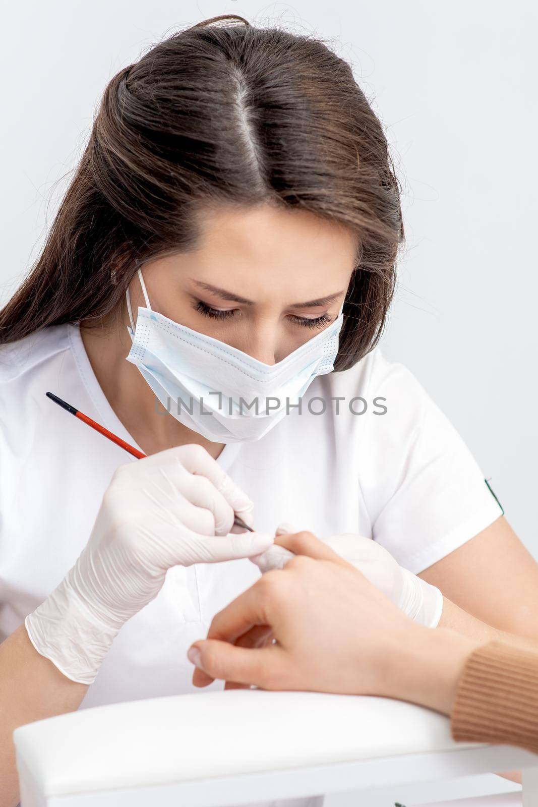 Manicure master with protective mask applying transparent nail polish on female nails in beauty salon