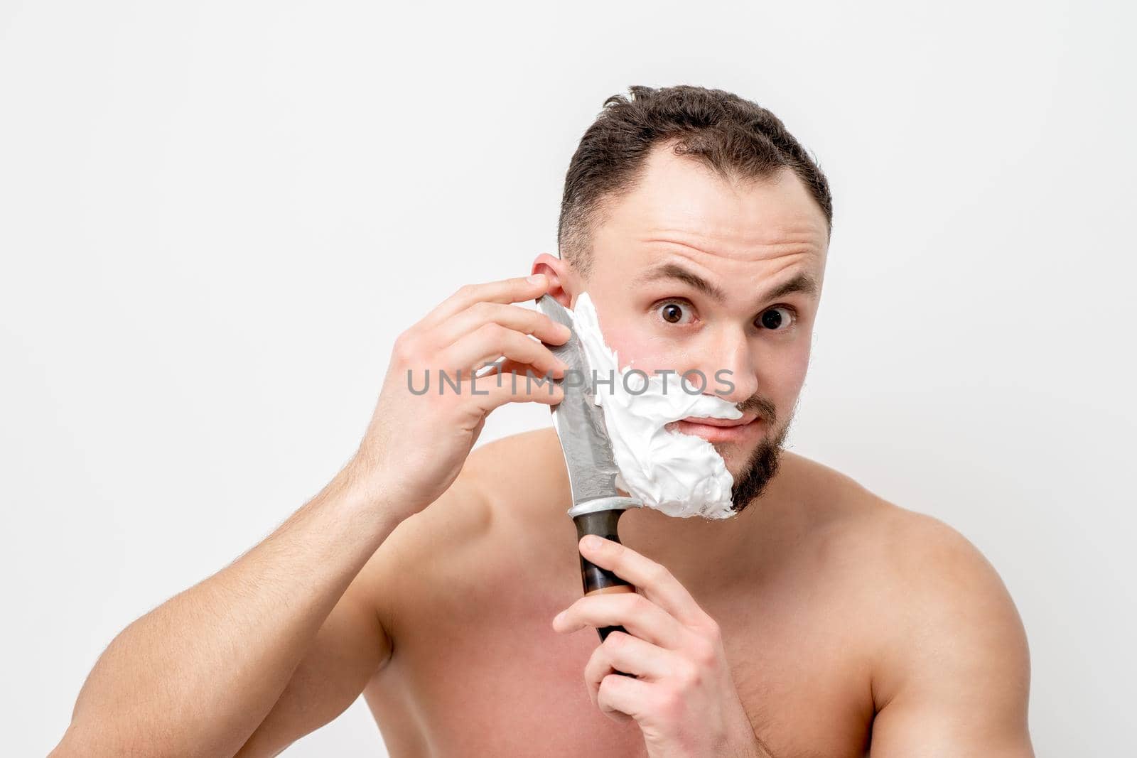 Young caucasian man shaving beard with knife on white background