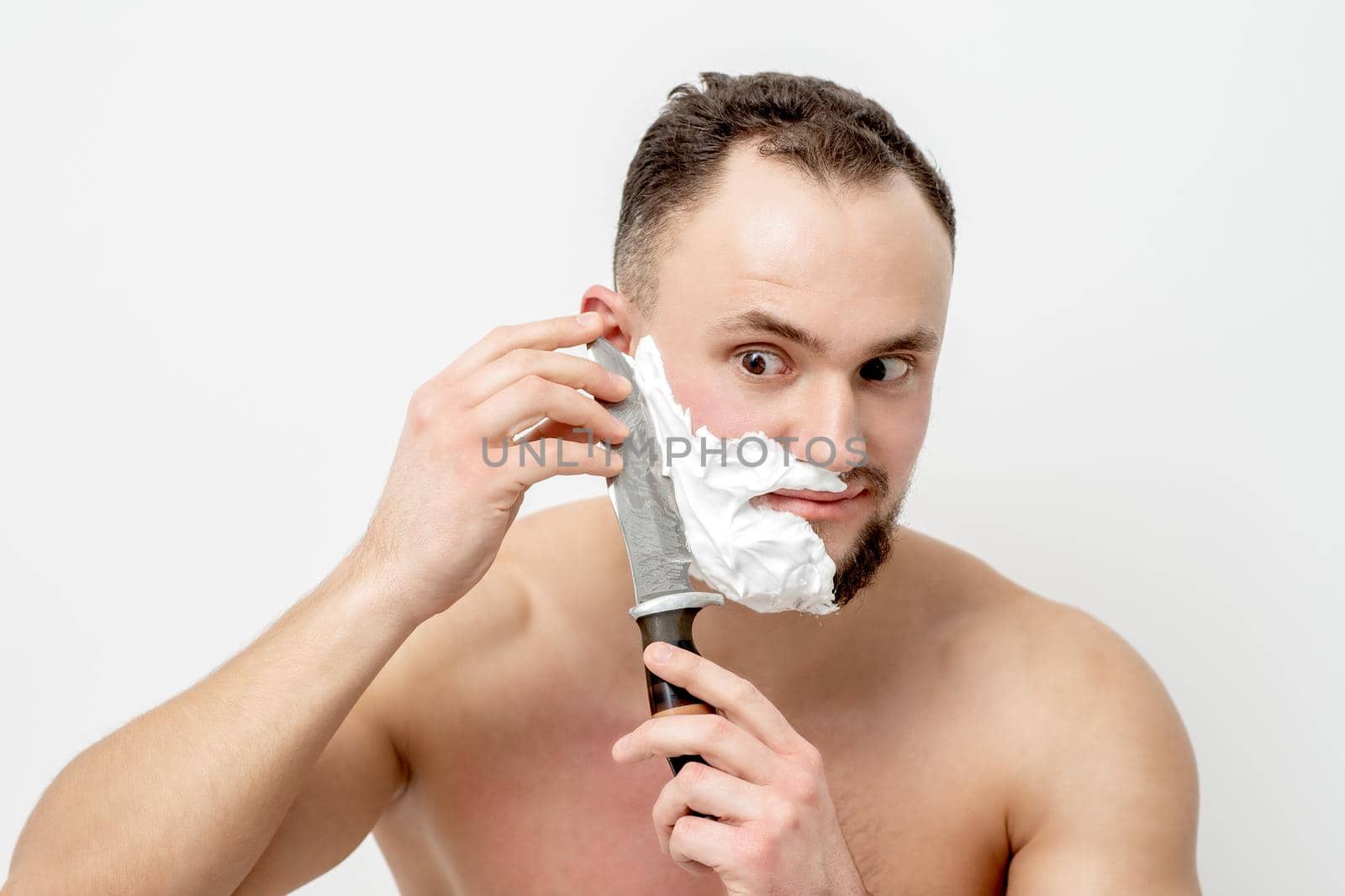 Young caucasian man shaving beard with knife on white background