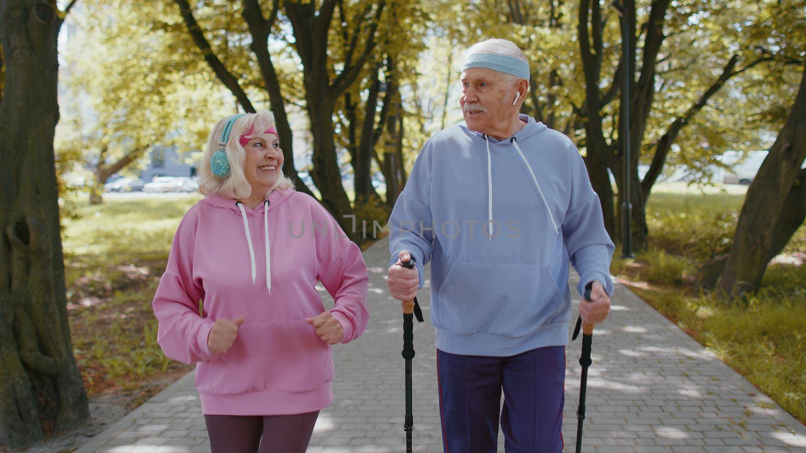 Active senior elderly couple 80 years old in summer park. Caucasian grandfather man training Nordic walking with ski trekking poles. Grandmother woman is running. Mature retired family sport hobby