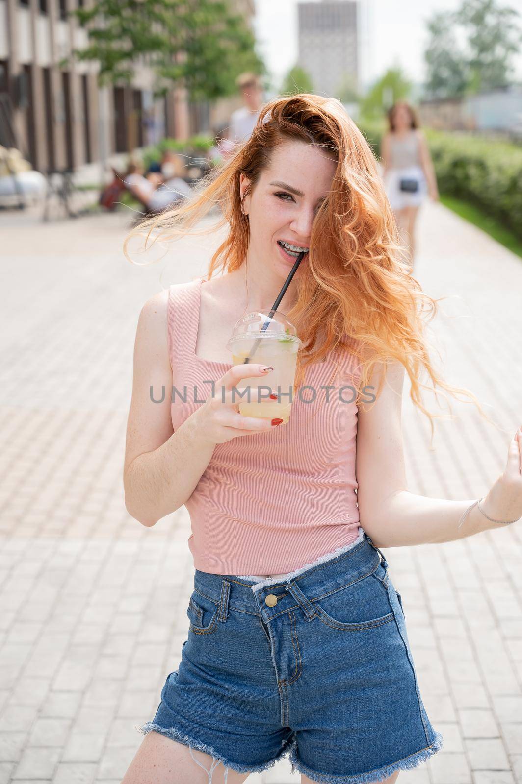 Young beautiful red-haired woman with braces drinks cooling cocktail outdoors in summer. Portrait of a smiling girl with freckles by mrwed54