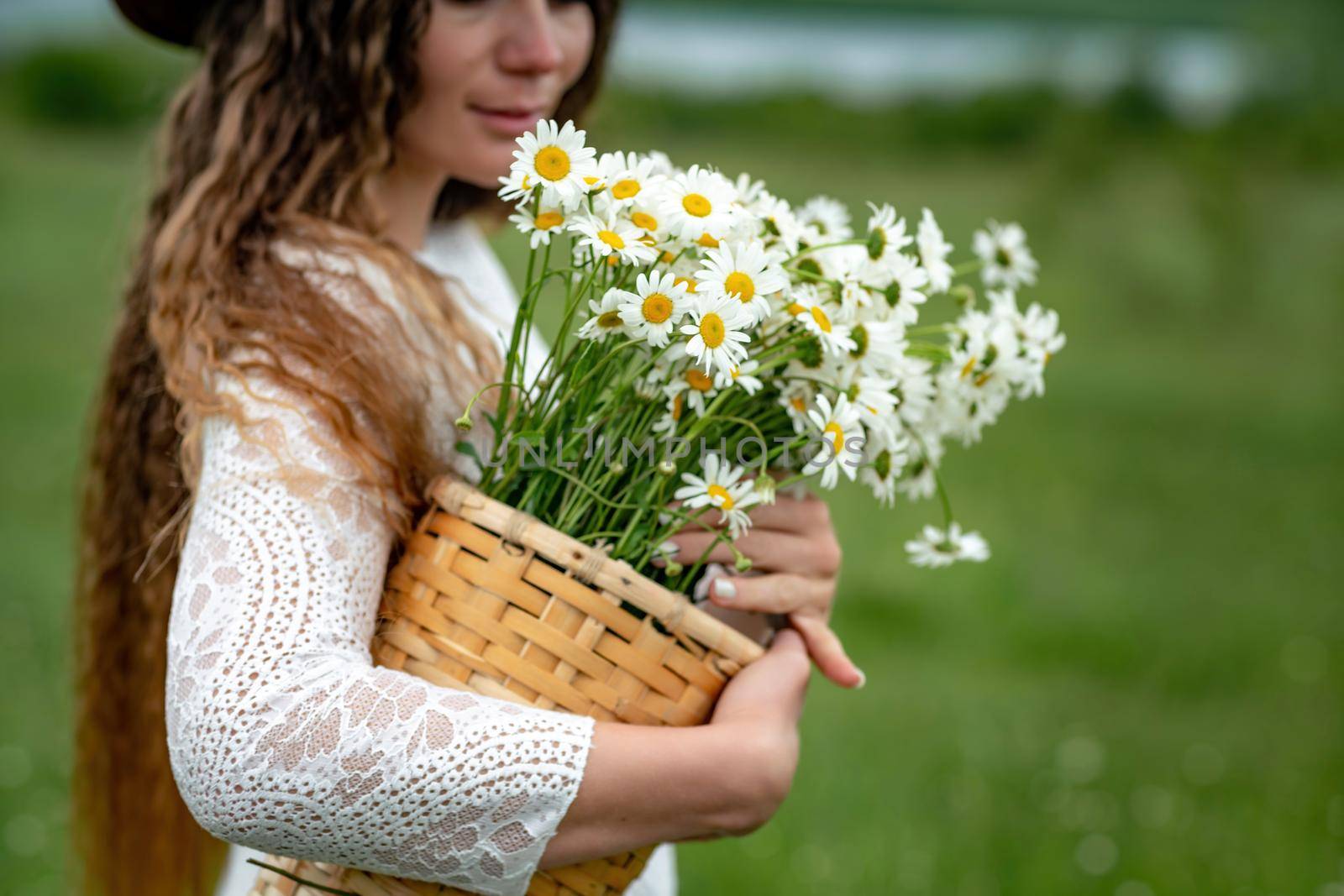 A middle-aged woman in a white dress and brown hat holds a lA middle-aged woman in a white dress and brown hat holds a basket in her hands with a large bouquet of daisies.arge bouquet of daisies in her hands. Wildflowers for congratulations.