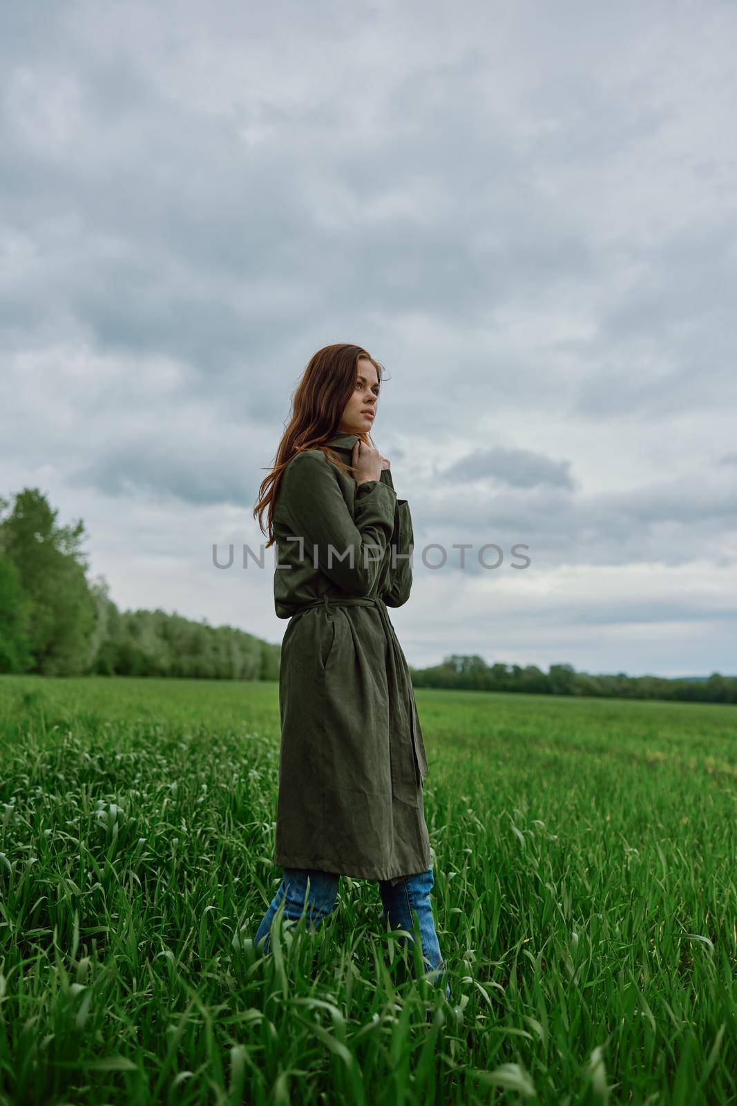 a beautiful woman in a dark coat stands in a green field in the spring in rainy weather by Vichizh