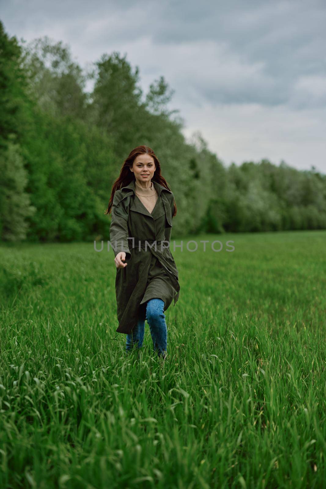 a beautiful woman in a long raincoat runs across a field in high grass in spring in cloudy weather. High quality photo