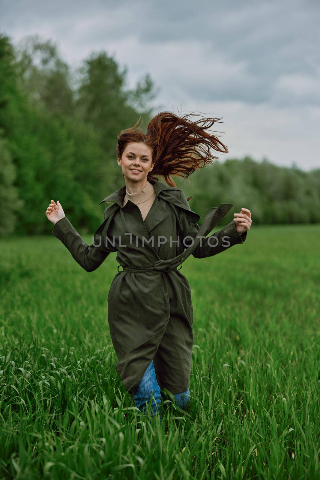 a beautiful woman in a long raincoat runs across a field in high grass in spring in cloudy weather. High quality photo