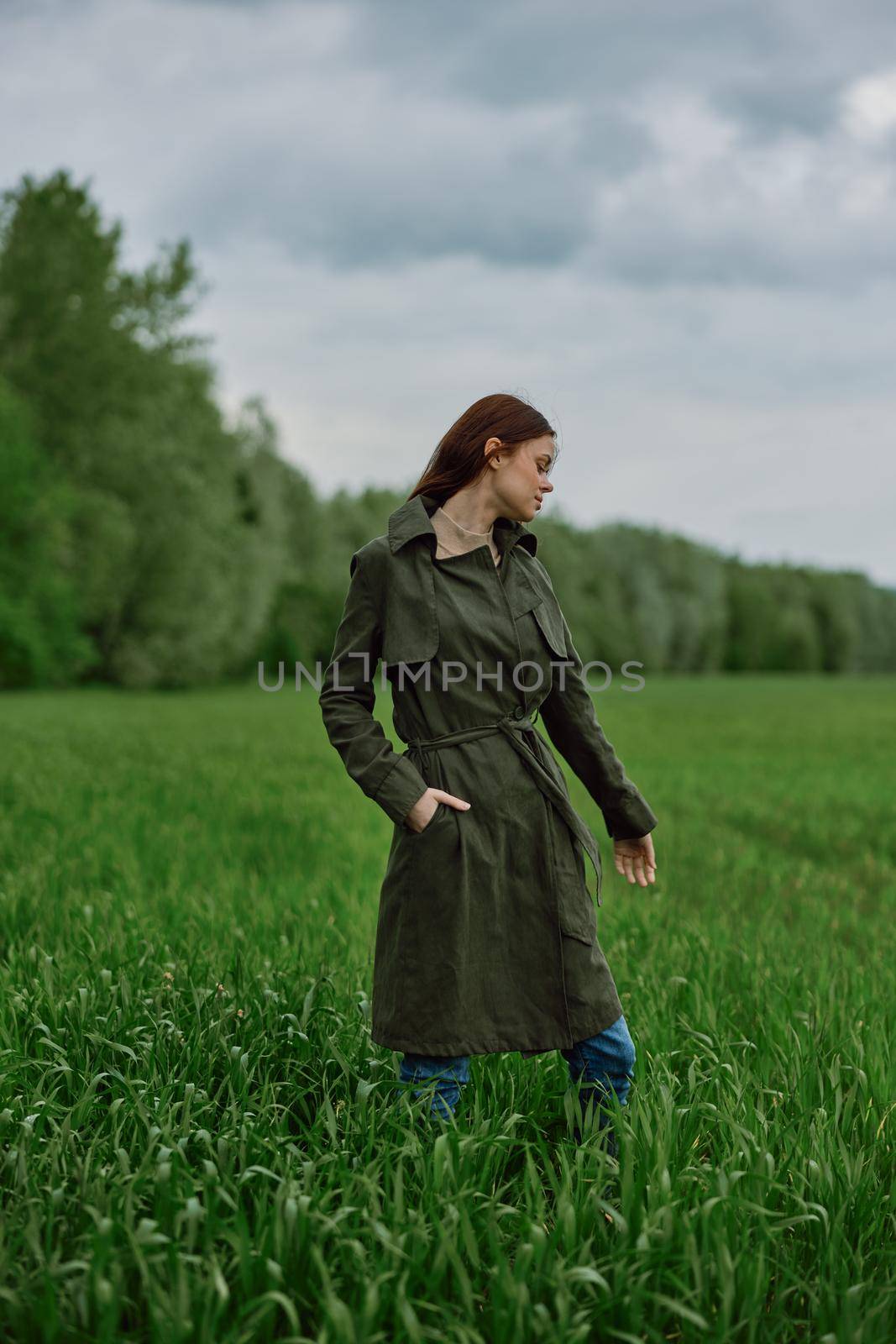 a woman in a long raincoat stands in tall green grass in a field in rainy weather in spring by Vichizh