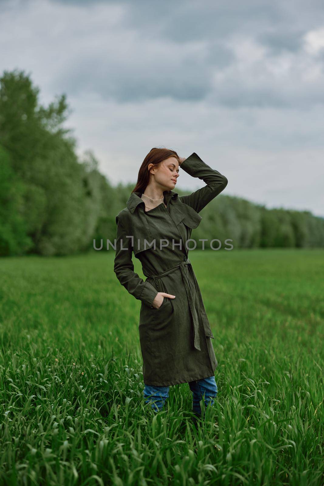 a beautiful woman in a dark coat stands in a green field in the spring in rainy weather and straightens her hair by Vichizh