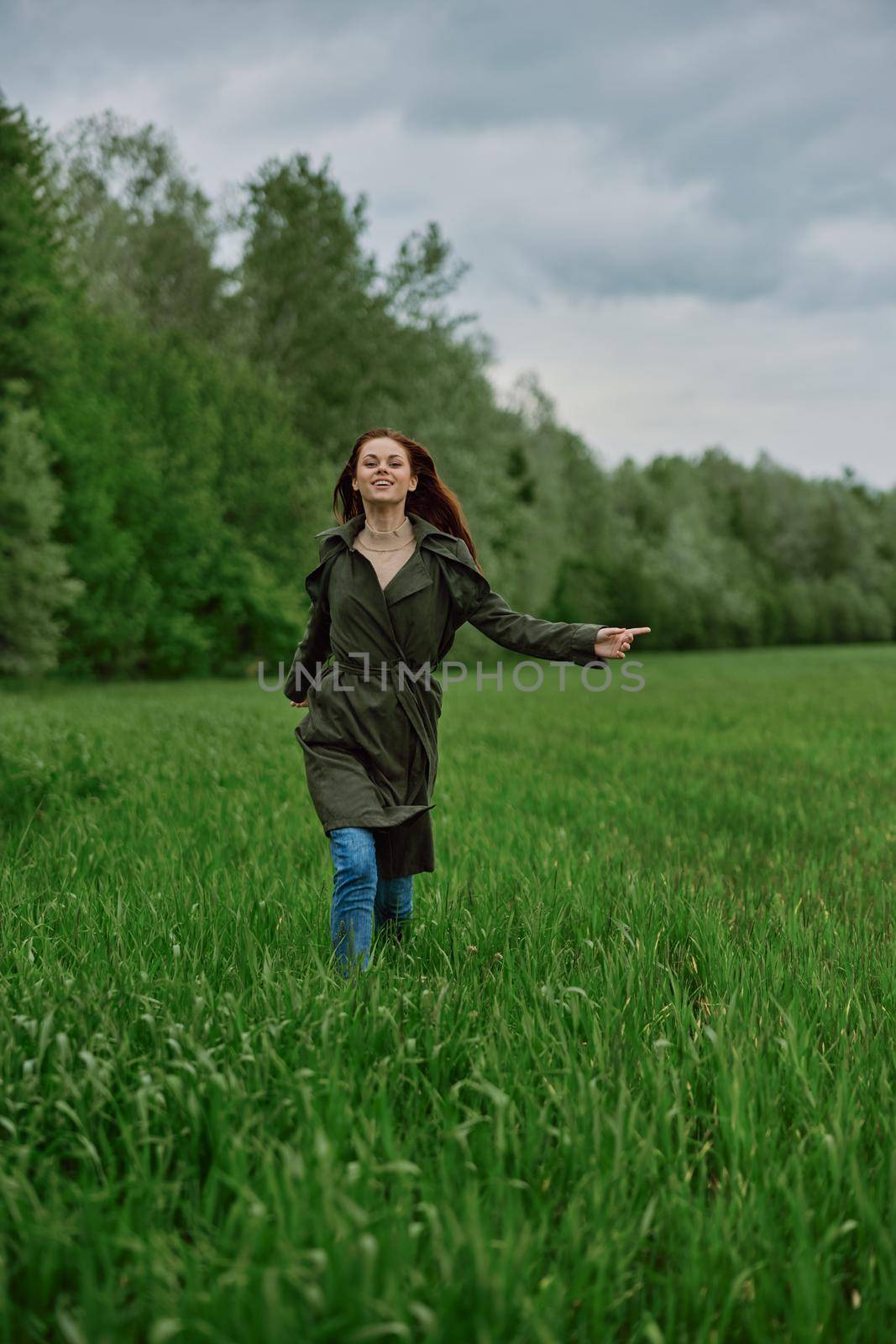 a beautiful woman in a long raincoat runs across a field in high grass in spring in cloudy weather by Vichizh