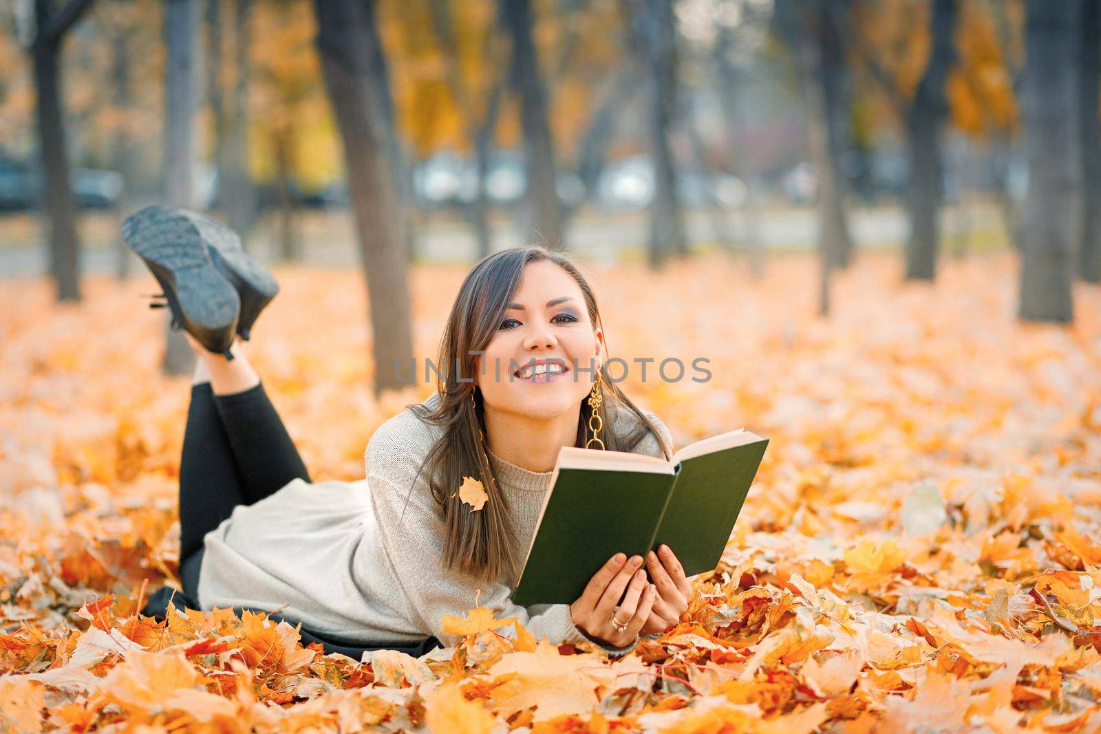 Young happy smiling woman lying on fall foliage and reading book in autumn park, look at camera.