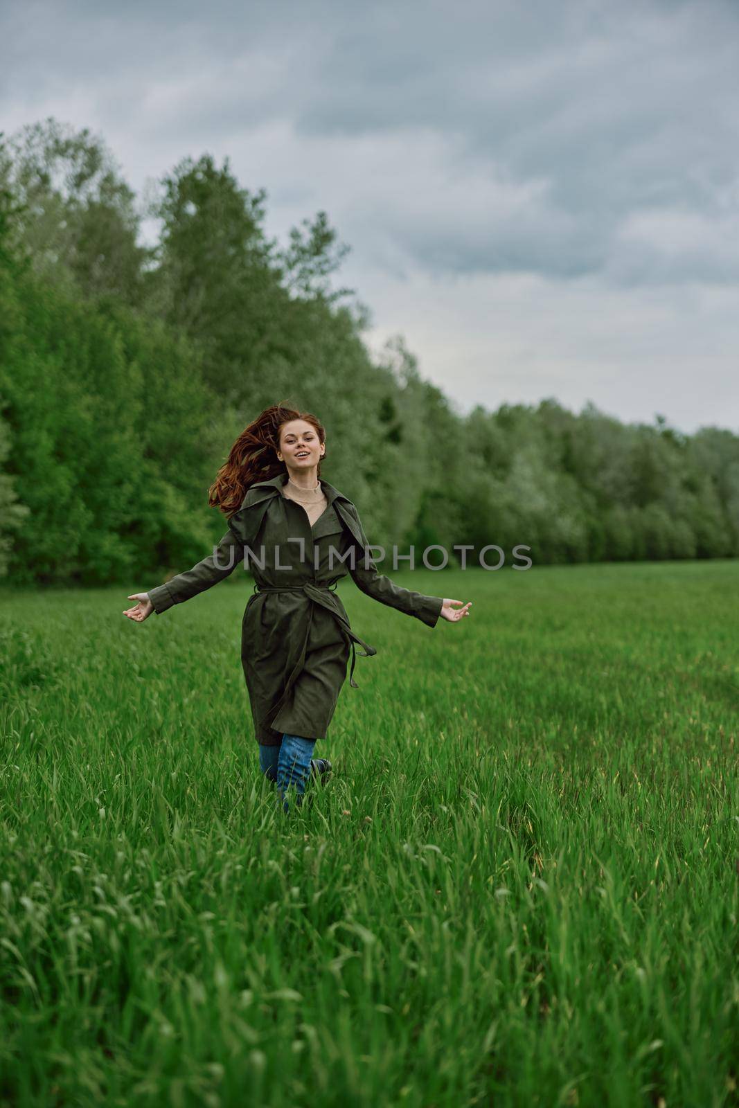 a beautiful woman in a long raincoat runs across a field in high grass in spring in cloudy weather. High quality photo