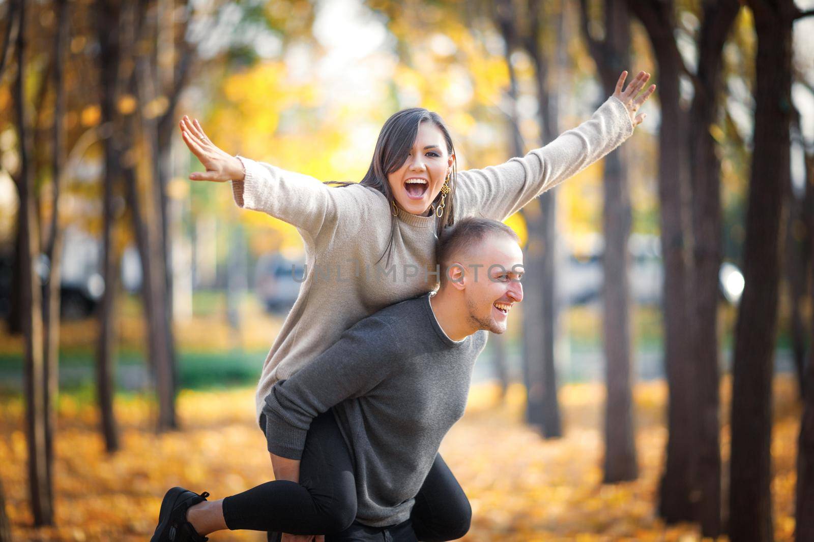 Happy smiling man carrying his laughing girlfriend on back, her opened arms like wings of an airplane having fun.