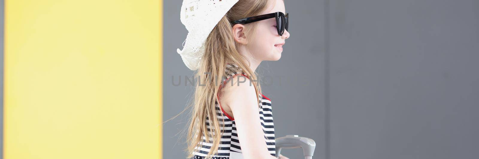 Portrait of well dressed little girl going on vacation carry yellow suitcase with her. Kid in cute hat and lovely summer dress. Airport, childhood concept