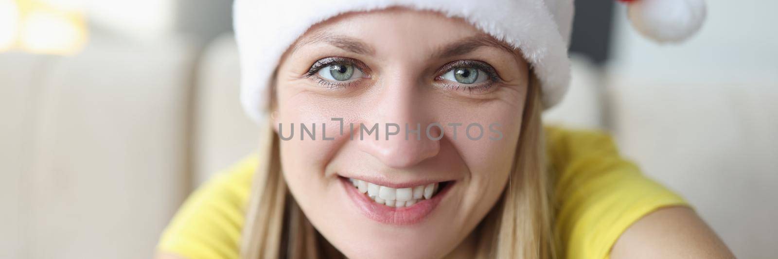 Portrait of beautiful blonde young woman posing on camera in santa claus hat and smile. Celebrating new year alone at home. Holiday, christmas mood concept