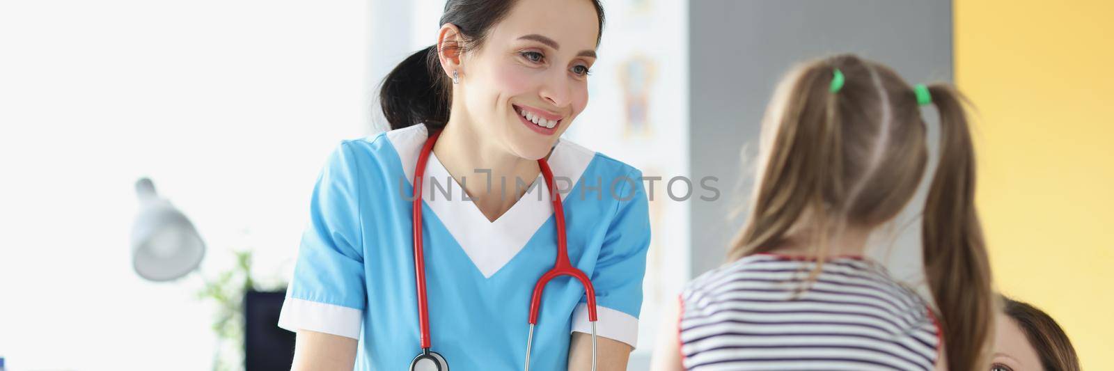Portrait of smiling professional pediatrician talk to young cute patient girl on appointment. Mum support daughter on her visit to doctor. Health concept