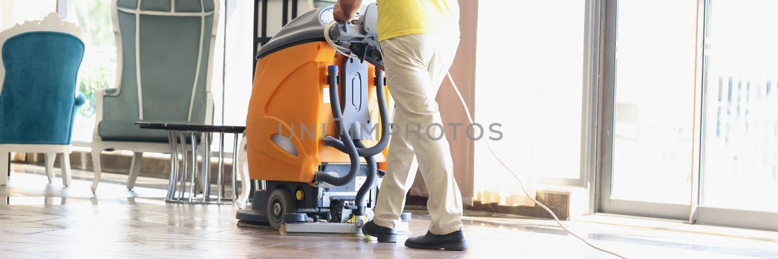Close-up of male cleaner cleaning interior of modern hotel lobby. Man in uniform with floor washer machine. Janitor in hall using washing vacuum cleaner. Cleaning service concept