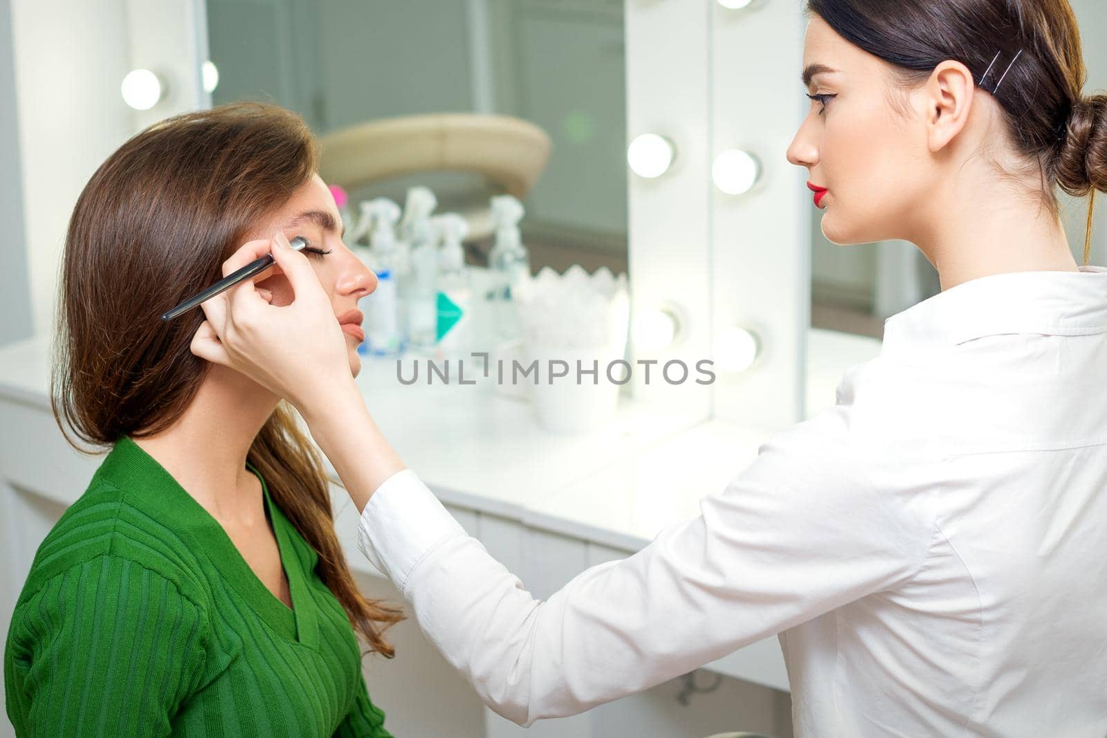 Makeup artist applies eyeshadow powder on eyes of young caucasian woman by brush tool in beauty salon