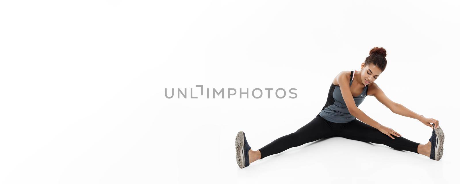 Sport, training, lifestyle and Fitness concept - portrait of beautiful happy African American woman stretching leg while sitting. Isolated on white studio background