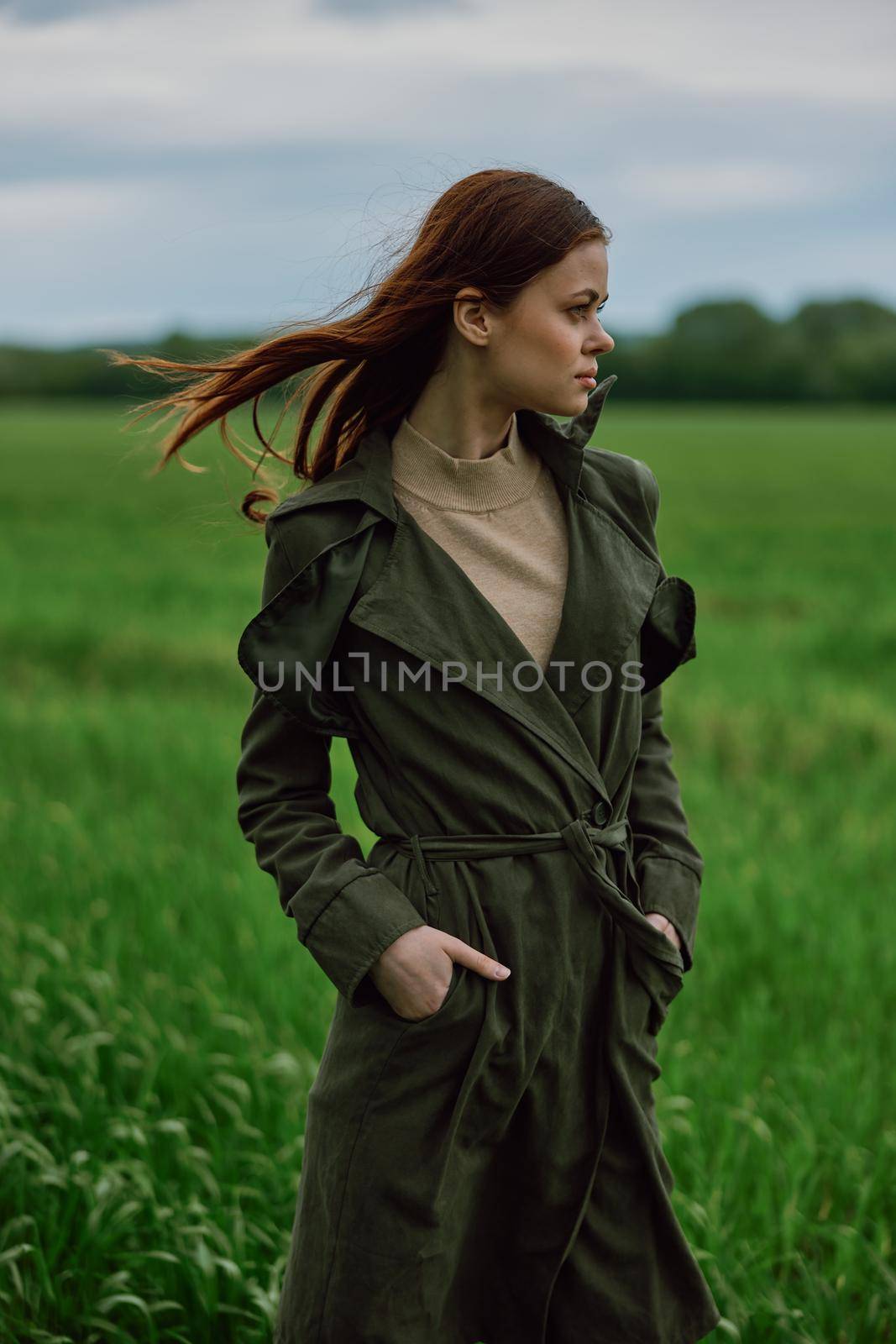 a beautiful red-haired woman stands in a field in a long dark raincoat, holding her hands in her pockets by Vichizh